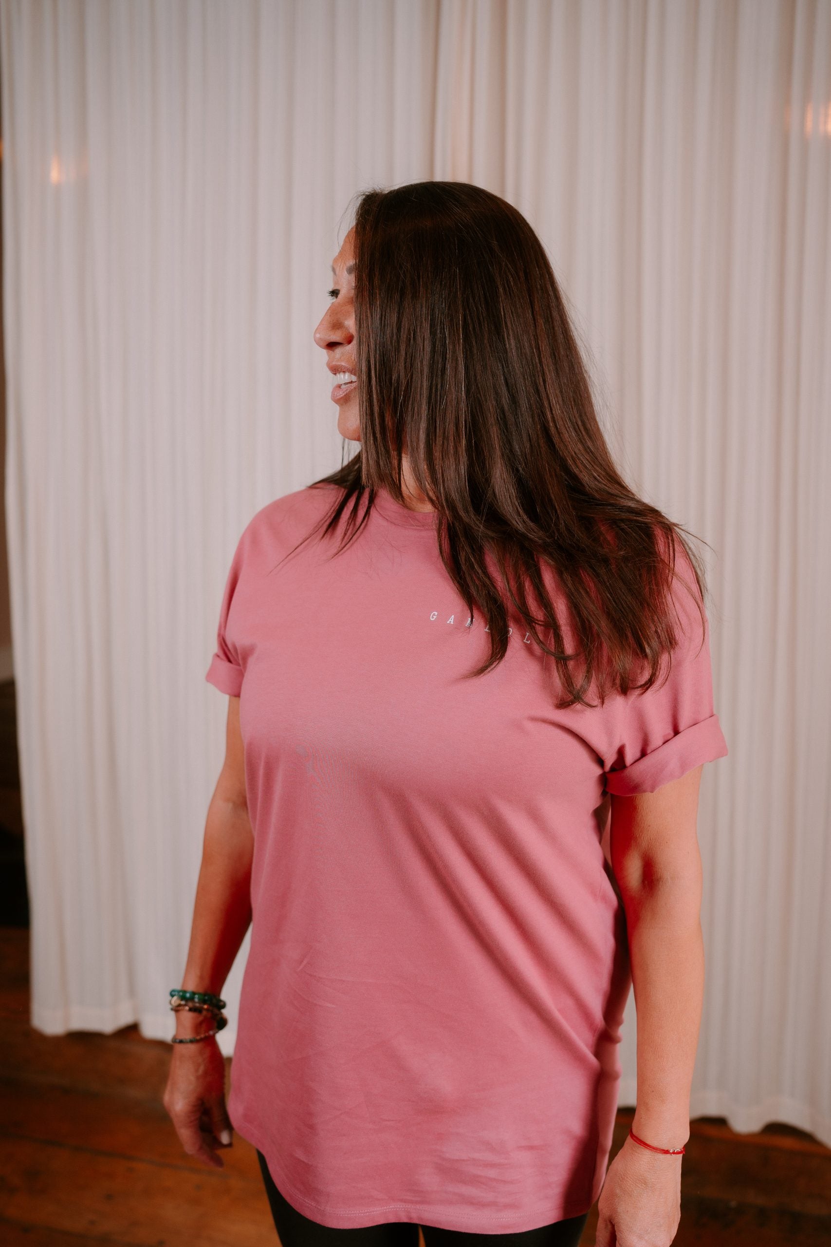 Woman wearing a pink t-shirt with a logo, standing against a white curtain.