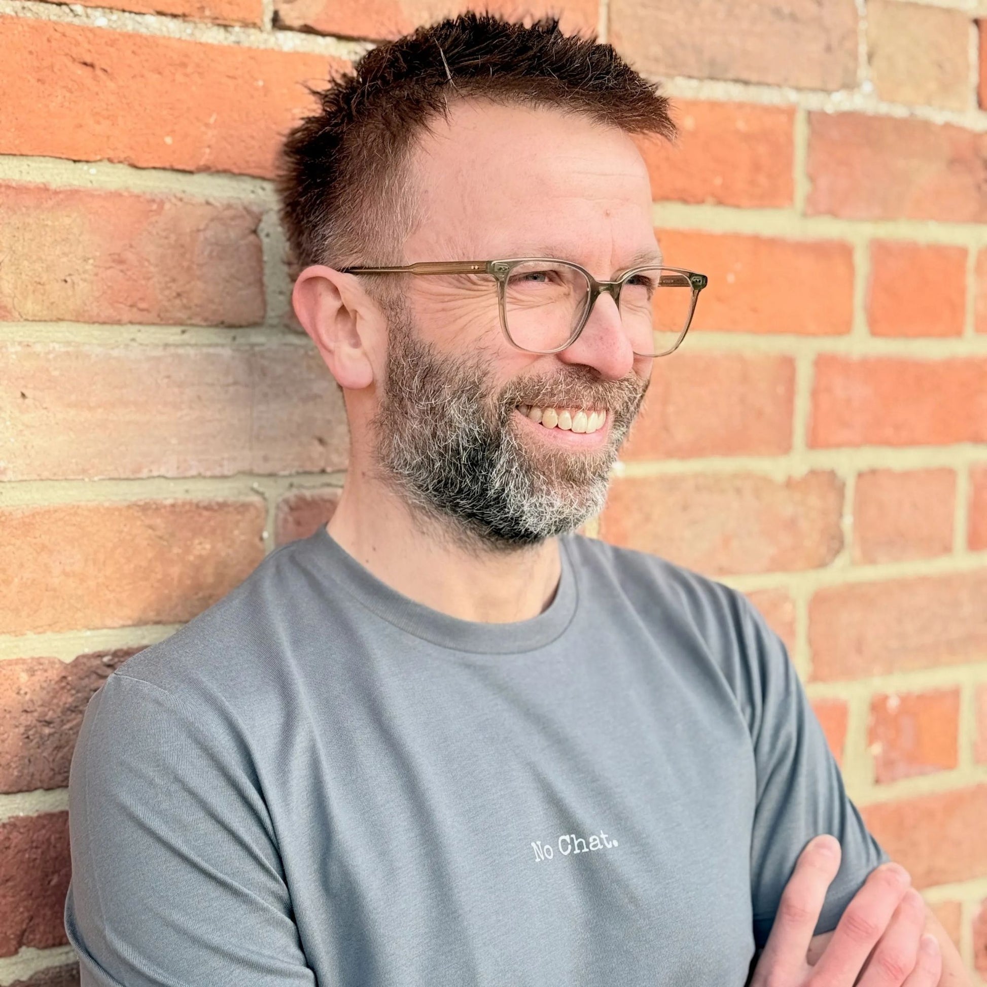 Man with glasses and a beard wearing a gray t-shirt against a brick wall.