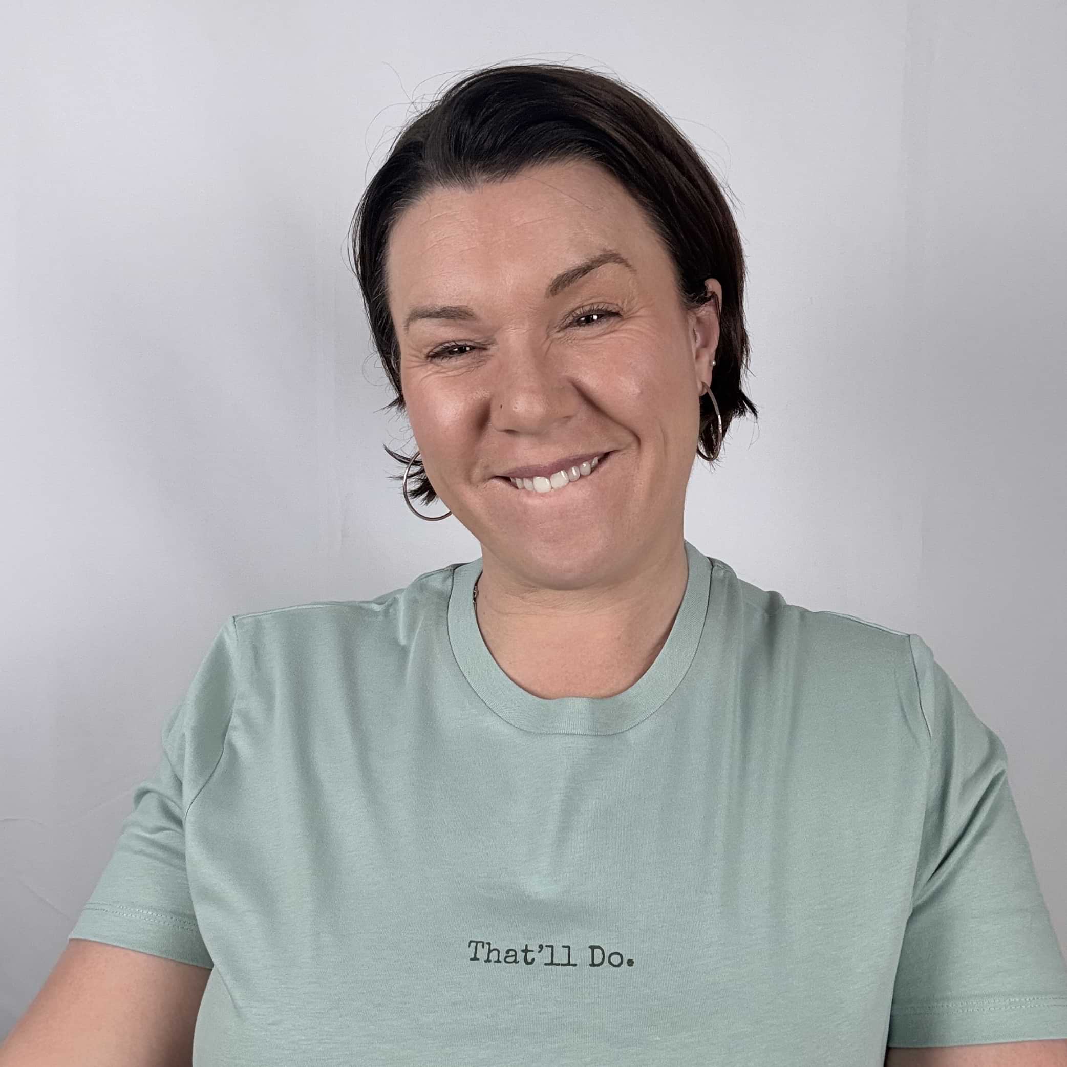Person wearing a light green t-shirt with 'That'll Do.' text on a plain background
