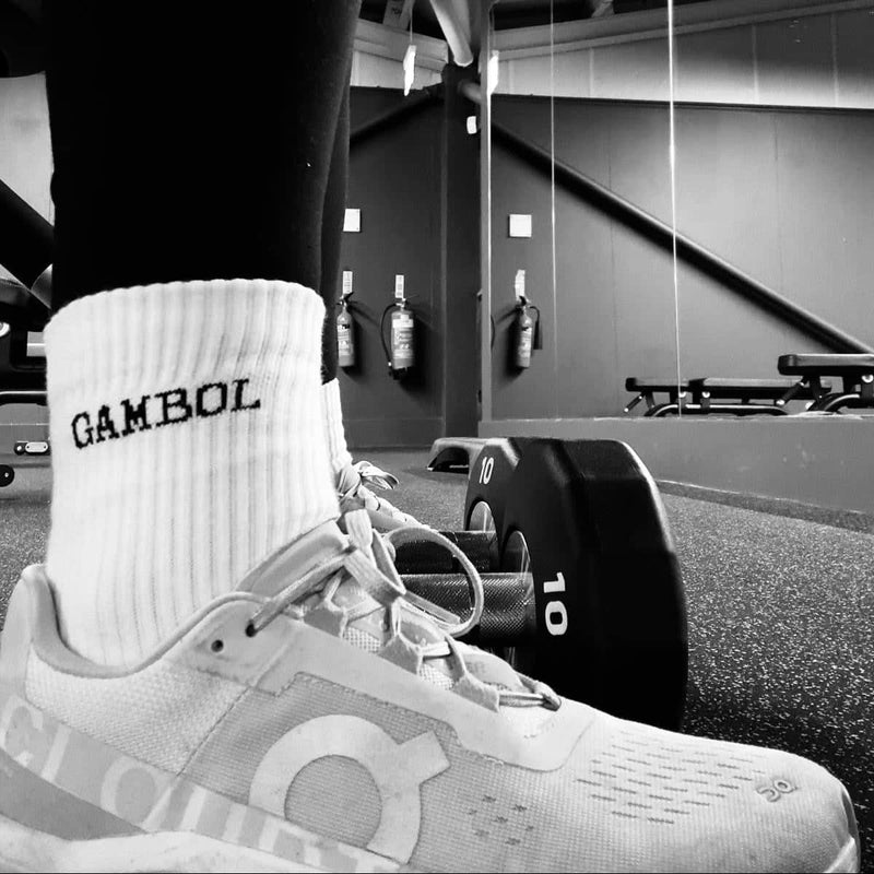 Person wearing a white sock with 'Gambol' branding and a gray athletic shoe in a gym setting.