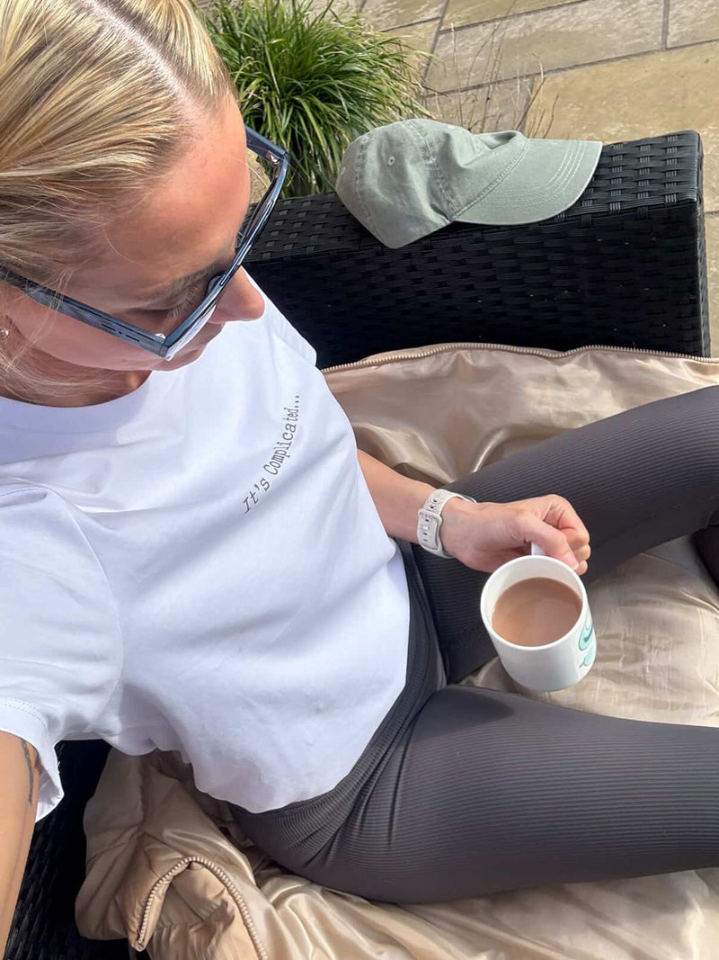 Woman sitting outdoors holding a cup, wearing glasses and a white t-shirt.
