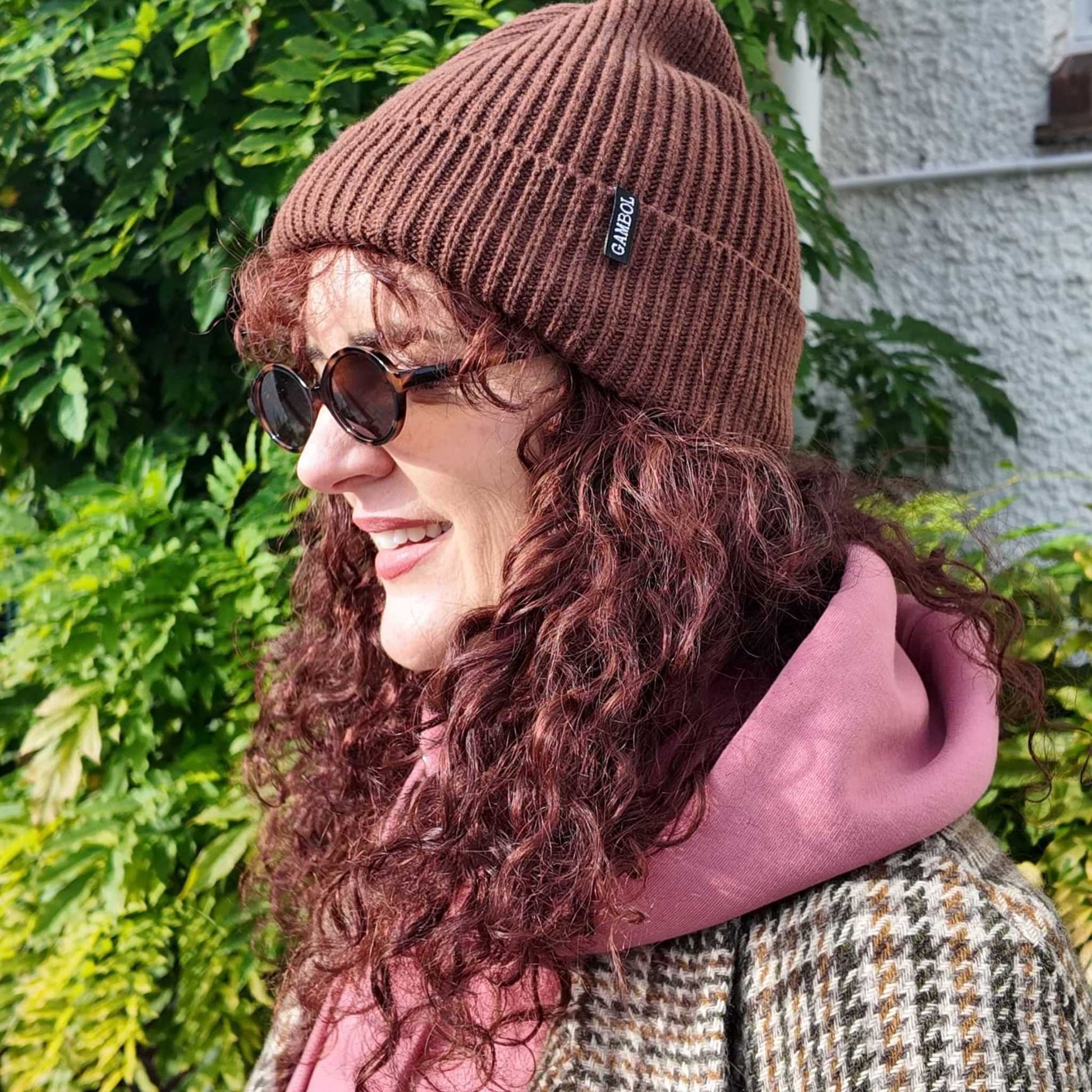 Woman wearing a brown beanie with a logo, sunglasses, and a pink hoodie outdoors.