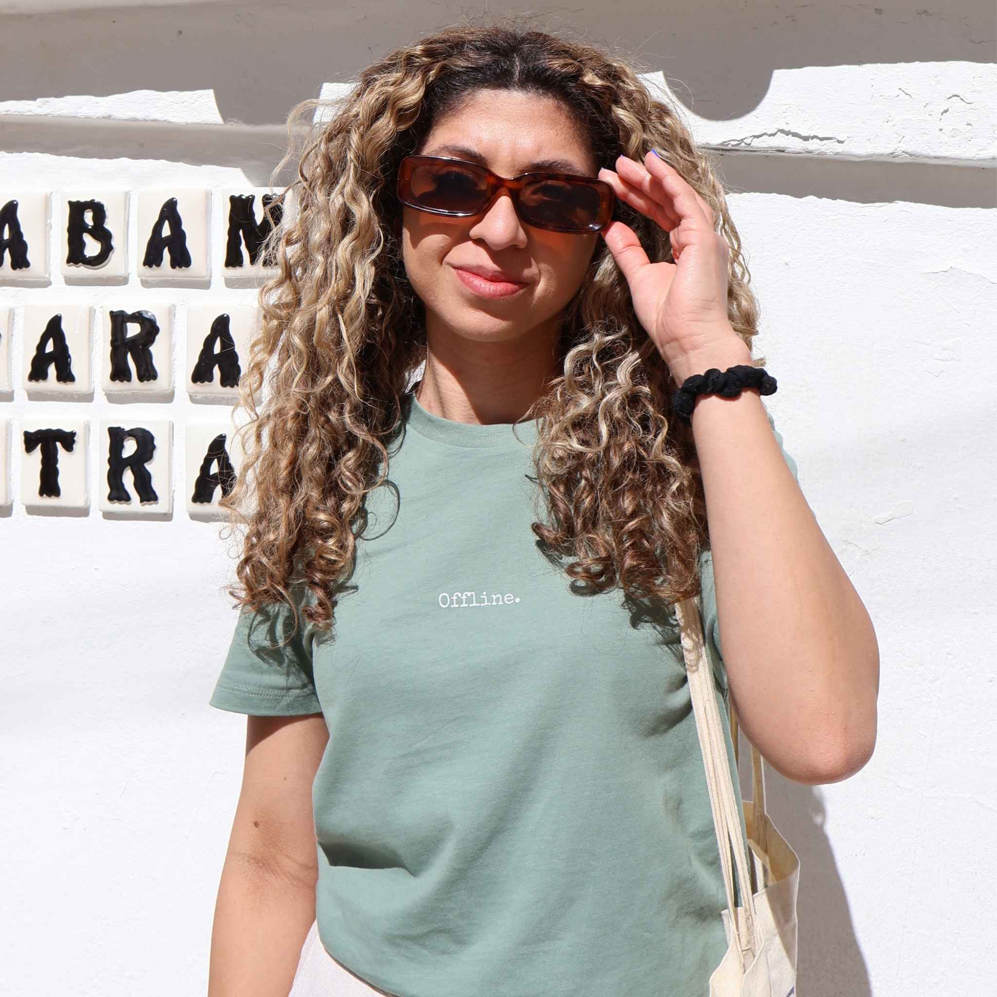 A woman with curly hair wearing a sage green t-shirt with 'OFFLINE.' printed on the chest, sunglasses, and a cream shoulder bag, standing against a white wall with decorative letter tiles.