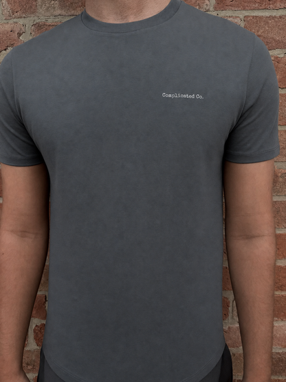 A man wearing a dark gray crewneck t-shirt with 'Complicated Co.' printed in white on the left chest, standing against a red brick wall.