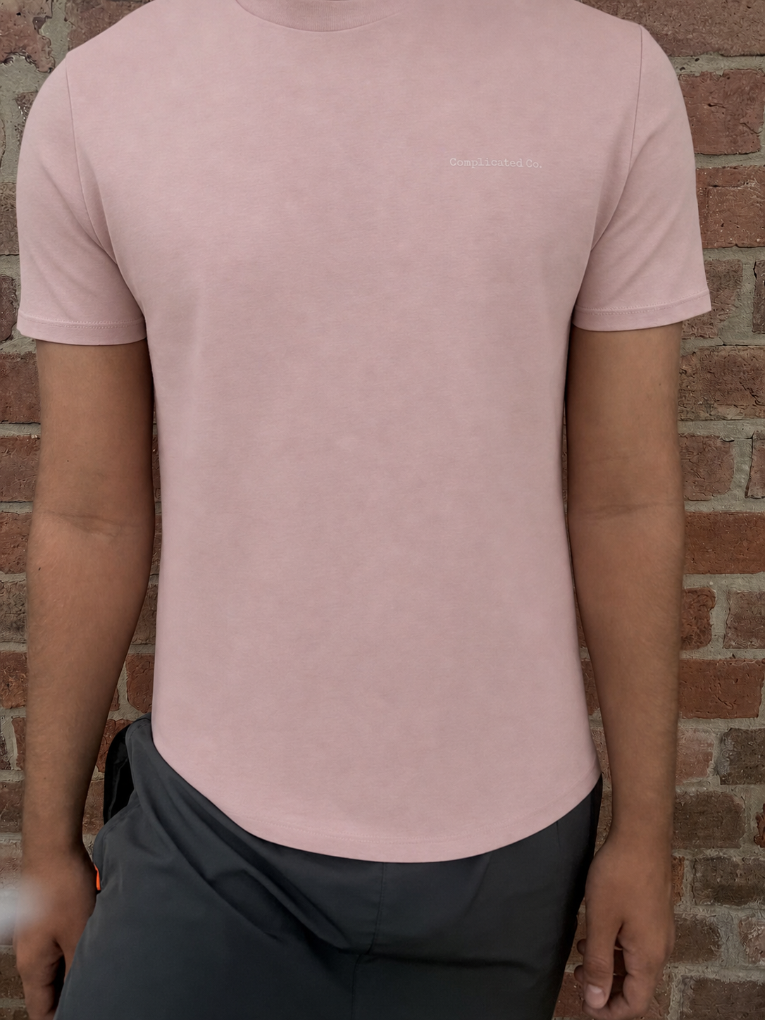 A man wearing a light pink short-sleeve crewneck t-shirt with a small white embroidered logo on the left chest, paired with dark gray pants, standing in front of a red brick wall.