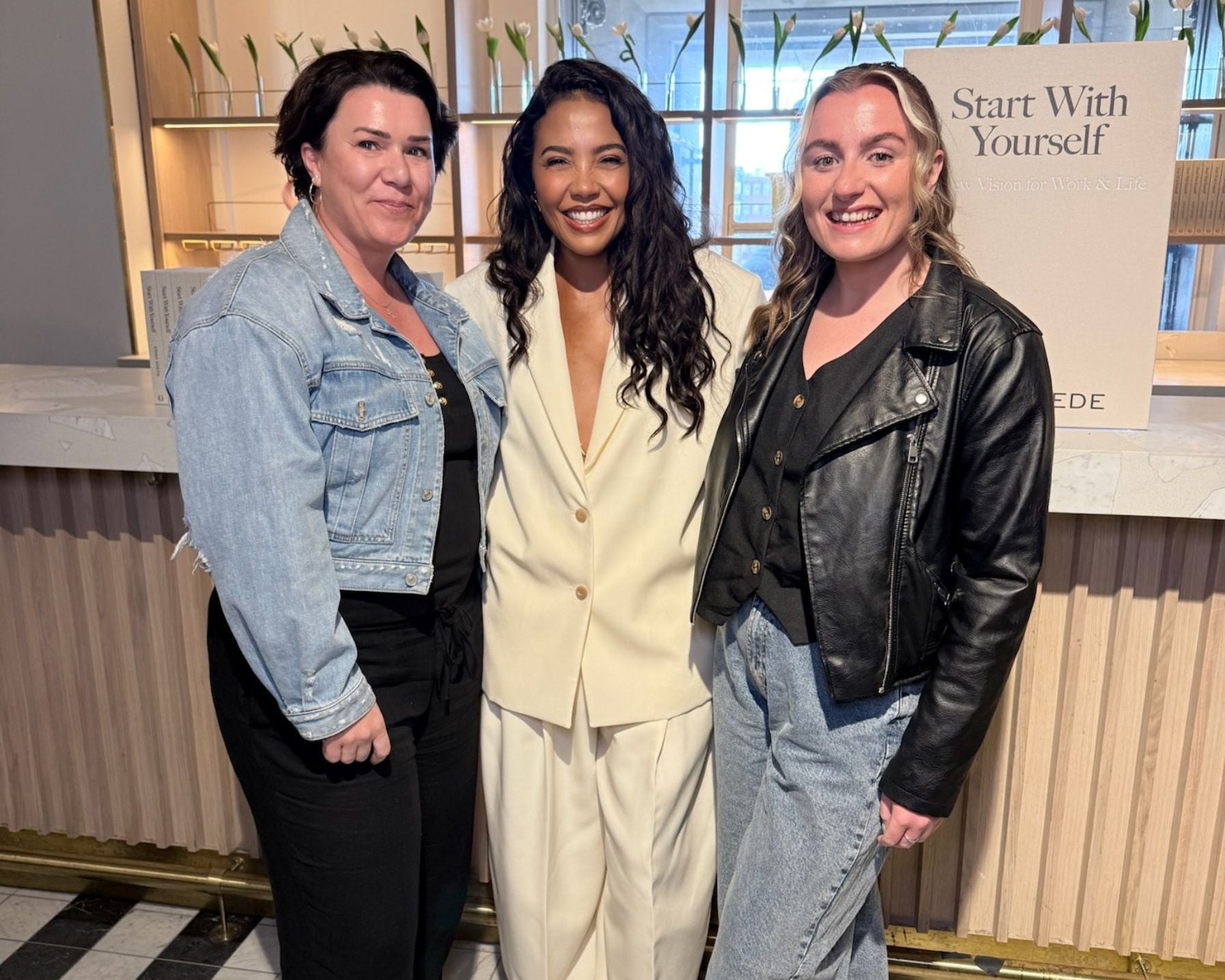 Three women posing together in a modern indoor setting with a sign in the background.