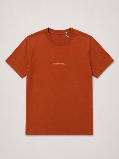 A rust-orange crewneck t-shirt with the word 'Parasocial.' printed in small white text on the left chest, displayed flat against a light gray background.
