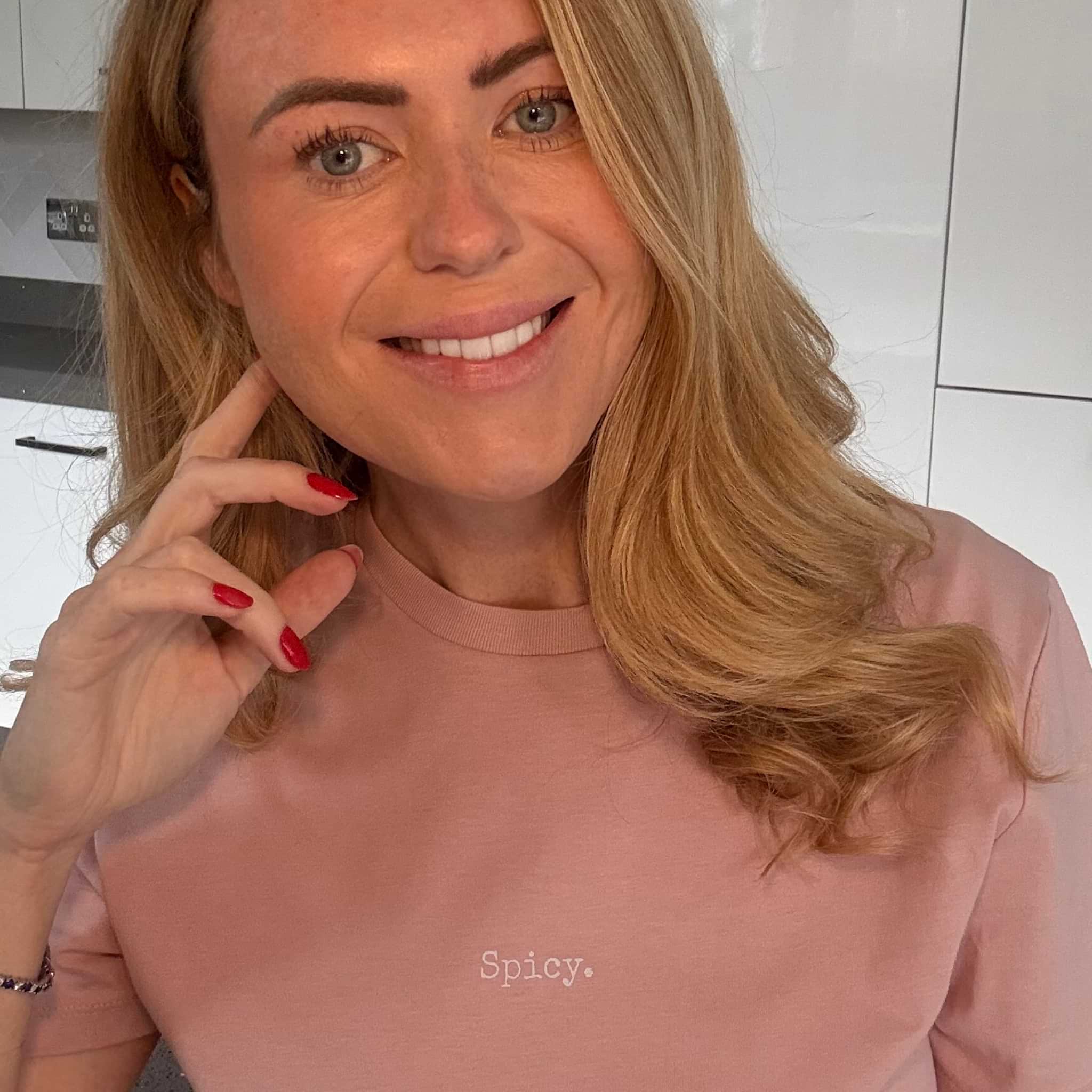Woman with blonde hair and red nails wearing a pink top in a kitchen setting.
